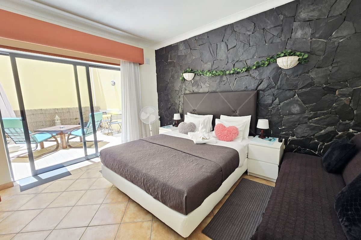 A bedroom with a stone wall, a bed with a brown headboard, a nightstand with a lamp, and a view of the outside patio.