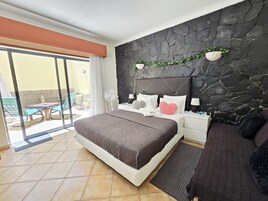 A bedroom with a stone wall, a bed with a brown headboard, a nightstand with a lamp, and a view of the outside patio.