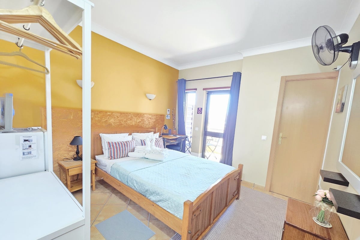 A bedroom with a wooden bed, a desk with a computer, a fan, and a door.