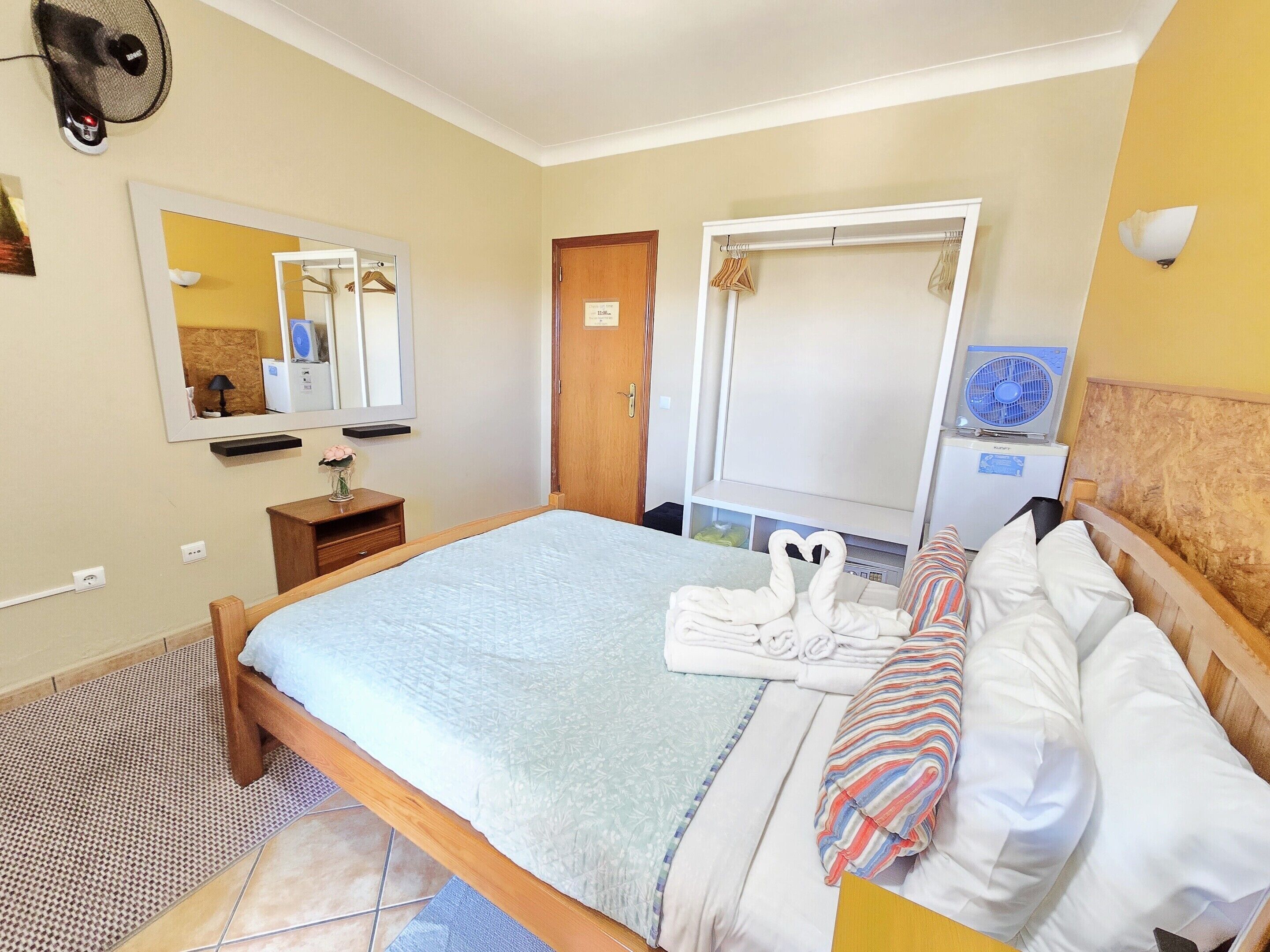A neatly arranged bedroom with a bed, bedside table, mirror, wardrobe, and a small desk with a lamp.