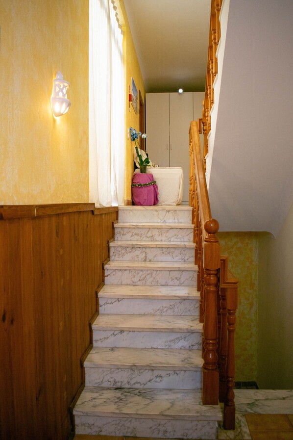 Un escalier avec des rampes en bois et des marches en marbre, menant à une pièce où se trouvent un lit et une valise rose, sur le palier.
