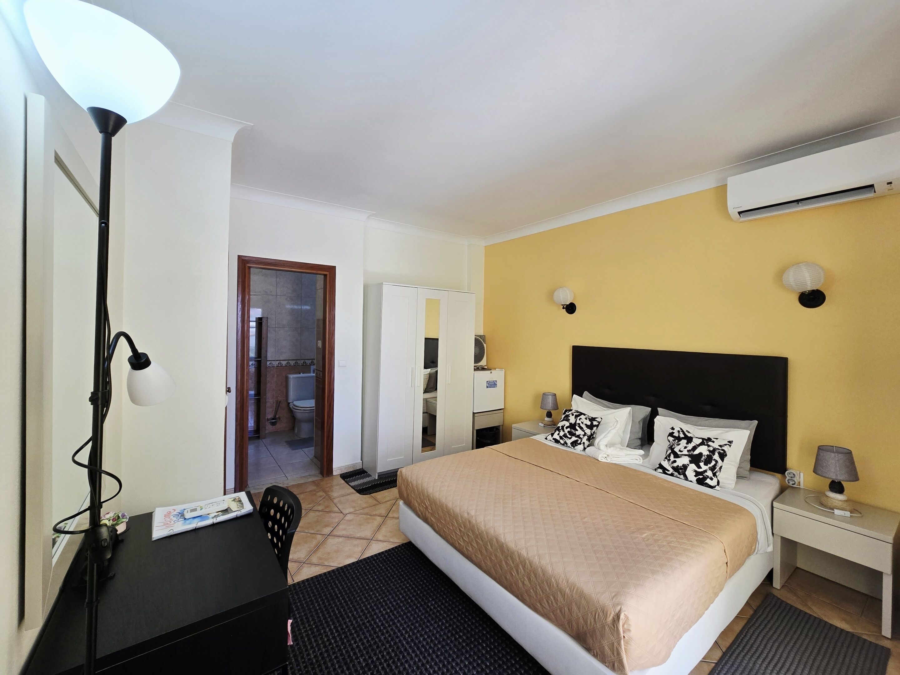 A hotel room with a bed, bedside tables, a desk, a chair, a lamp, an air conditioner, and a bathroom visible through an open door.