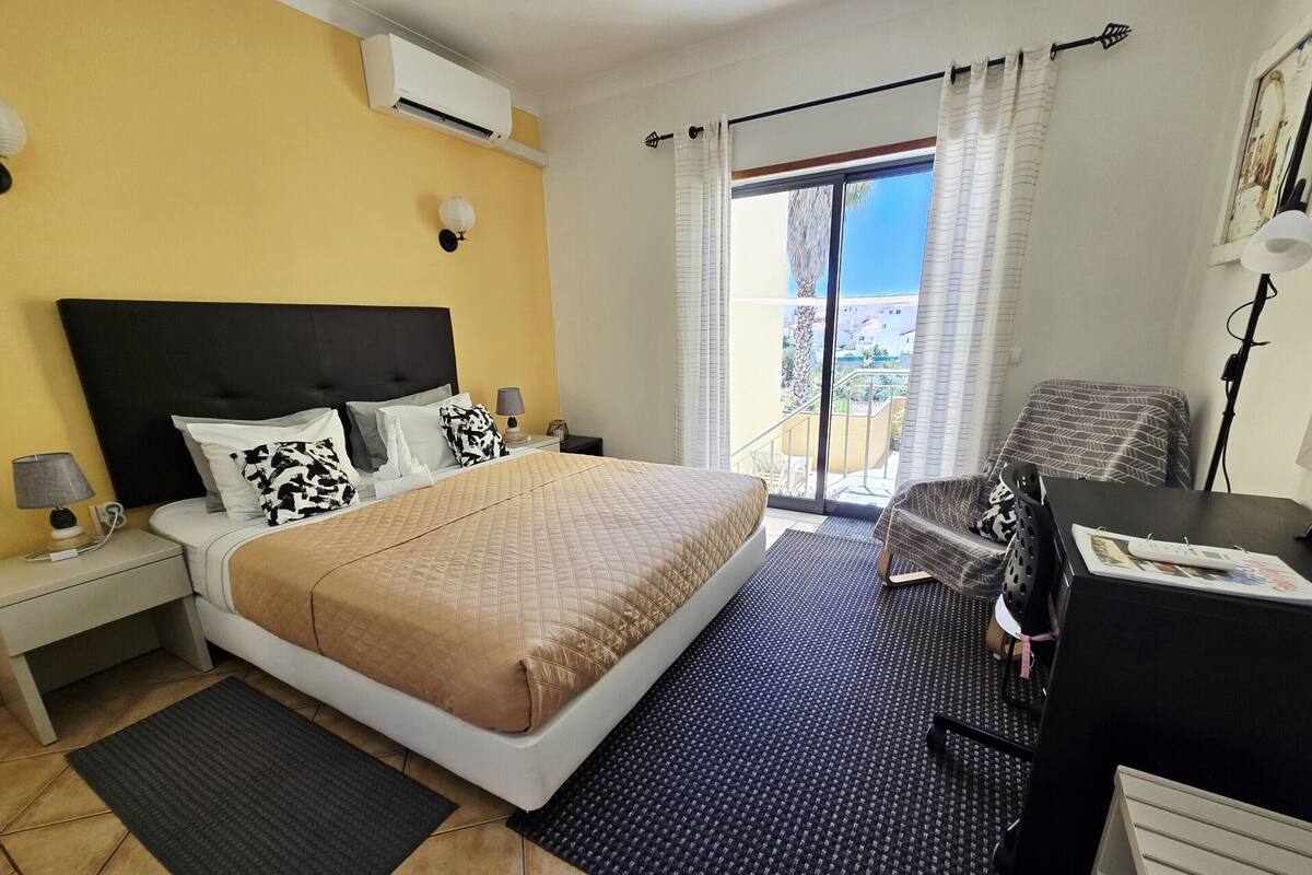 A hotel room with a bed, a desk, a chair, a balcony with a view, and an air conditioning unit.