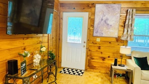 A cozy room with wooden walls, a television mounted on the wall, a white door, a checkered rug, a white sofa, and a table with a vase of flowers. - 3 bdrm 2 BA cabin with WiFi, AC near Surry, Hopewell, Wakefield& Prince George (Spring Grove)
