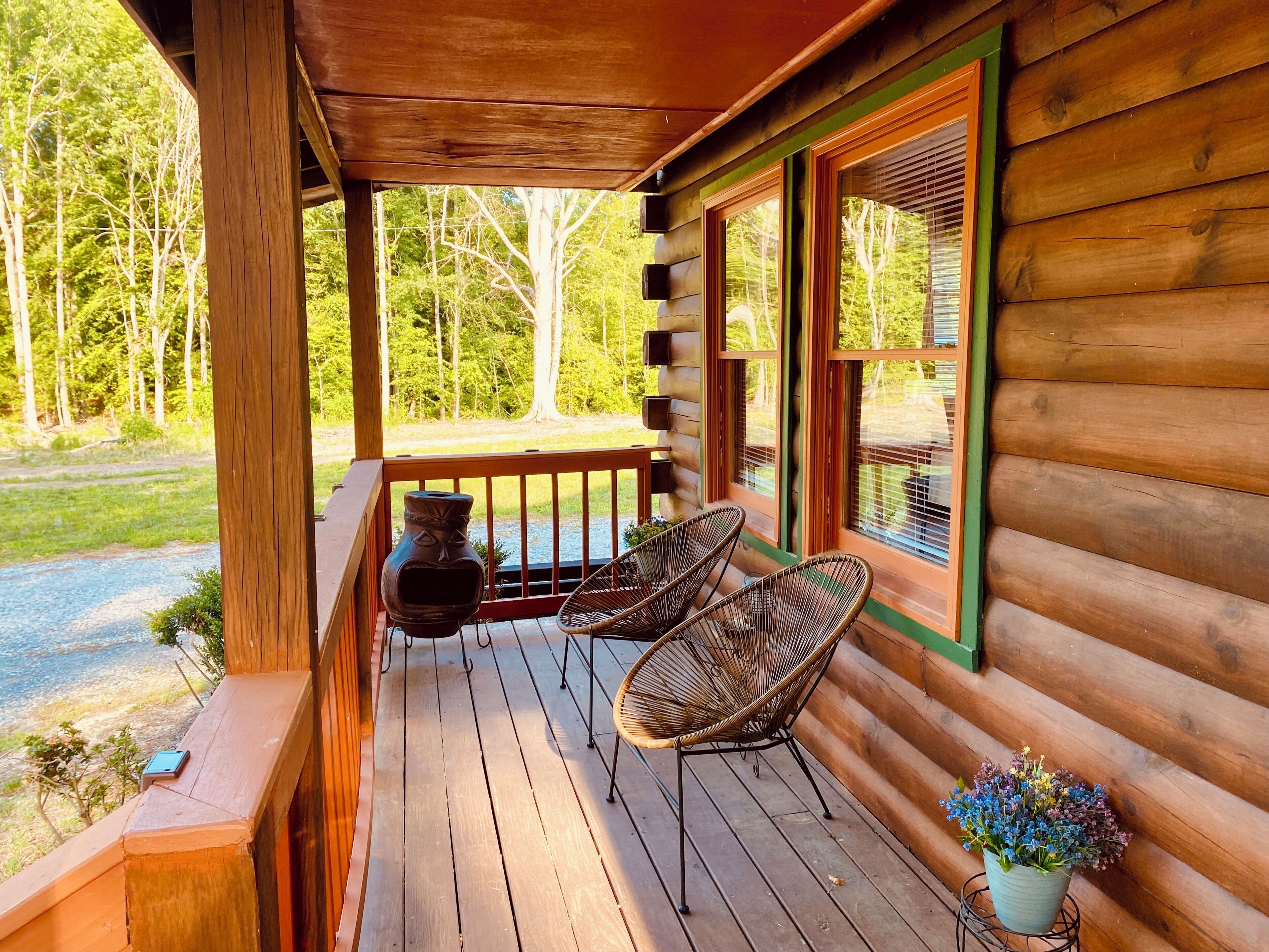 A wooden cabin porch with two wicker chairs, a potted plant, and a stone vase.