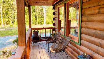 A wooden cabin porch with two wicker chairs, a potted plant, and a stone vase.