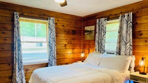 A wooden-paneled room with a bed, two bedside lamps, and windows with tree-patterned curtains. - 3 bdrm 2 BA cabin with WiFi, AC near Surry, Hopewell, Wakefield& Prince George (Spring Grove)