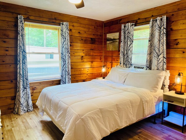 A wooden-paneled room with a bed, two bedside lamps, and windows with tree-patterned curtains. - 3 bdrm 2 BA cabin with WiFi, AC near Surry, Hopewell, Wakefield& Prince George (Spring Grove)