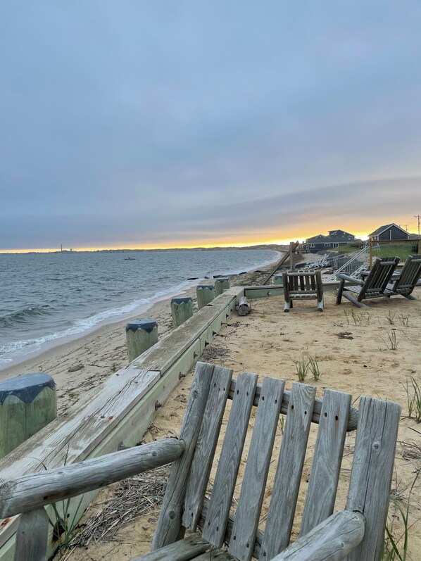 A wooden beach chair and a wooden fence along a sandy beach with a view of the ocean and distant houses.