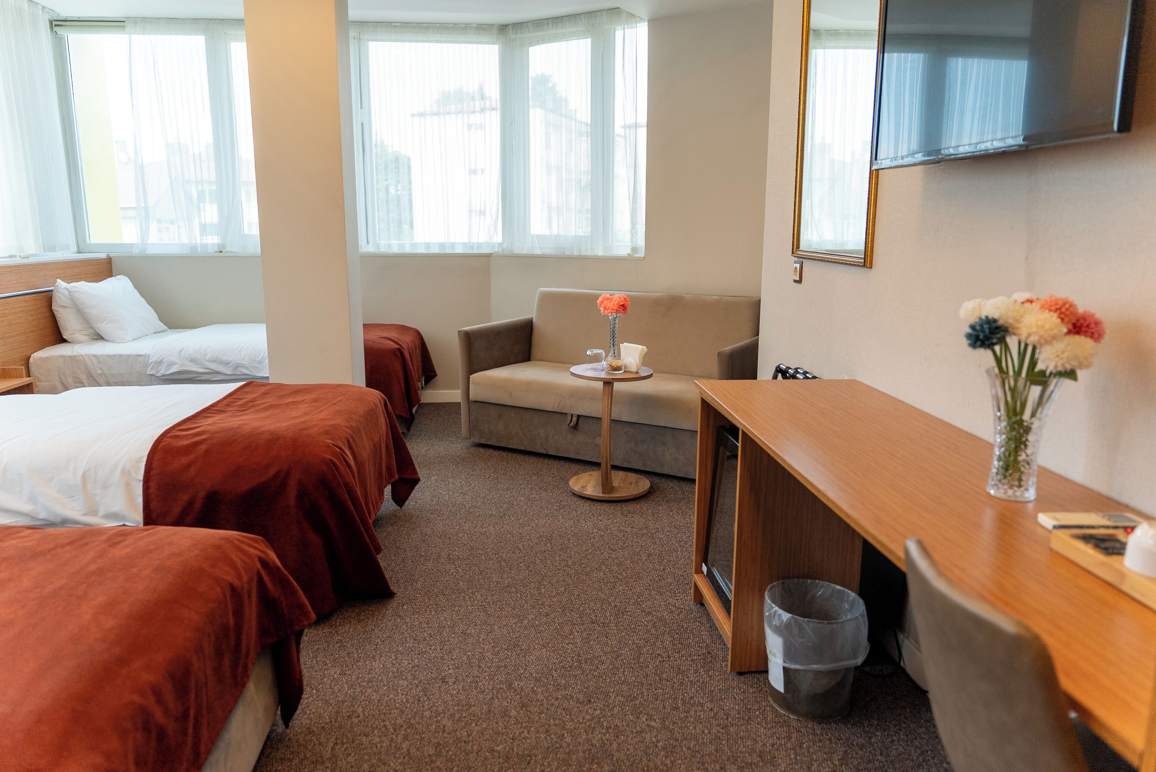 A hotel room with two beds, a sofa, a small table with a vase, a TV, and a trash bin.