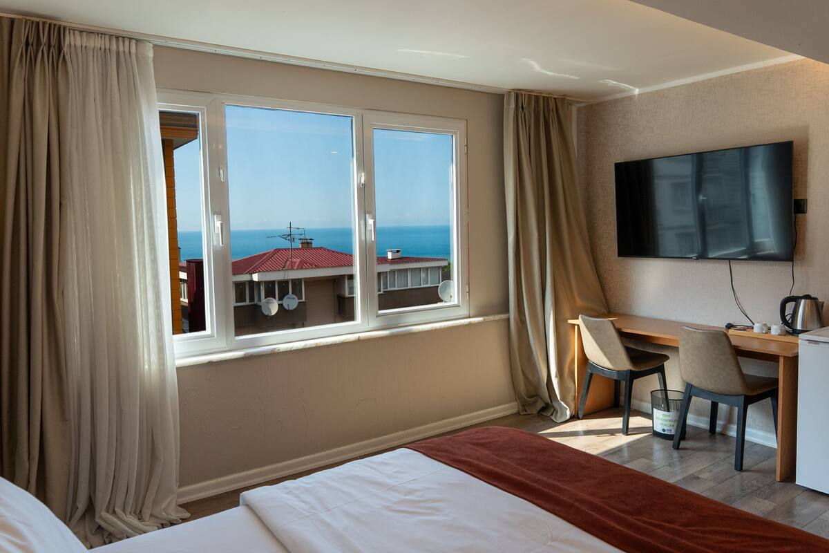 A hotel room with a bed, a desk, a chair, a television, and a window with a view of the sea.