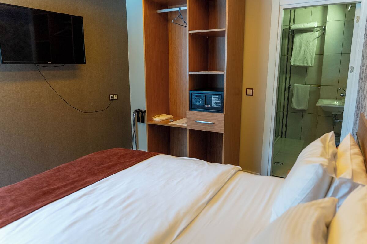 A hotel room with a bed, a TV mounted on the wall, a wooden wardrobe, a safe, and a bathroom visible through an open door.