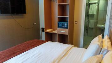 Habitación de hotel con cama, televisor montado en la pared, armario de madera, una caja fuerte y baño visible a través de una puerta abierta.