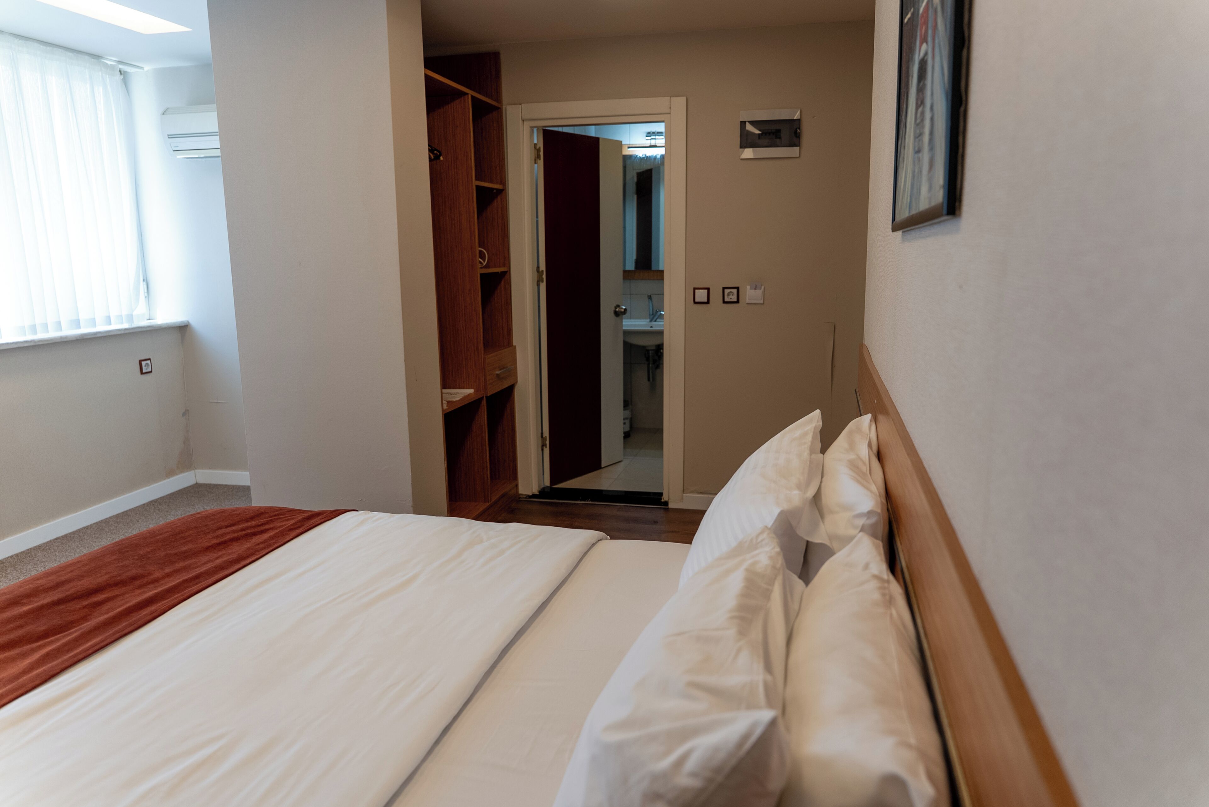Quarto de hotel com cama, armário de madeira, banheiro visível através de uma porta aberta e um quadro na parede.