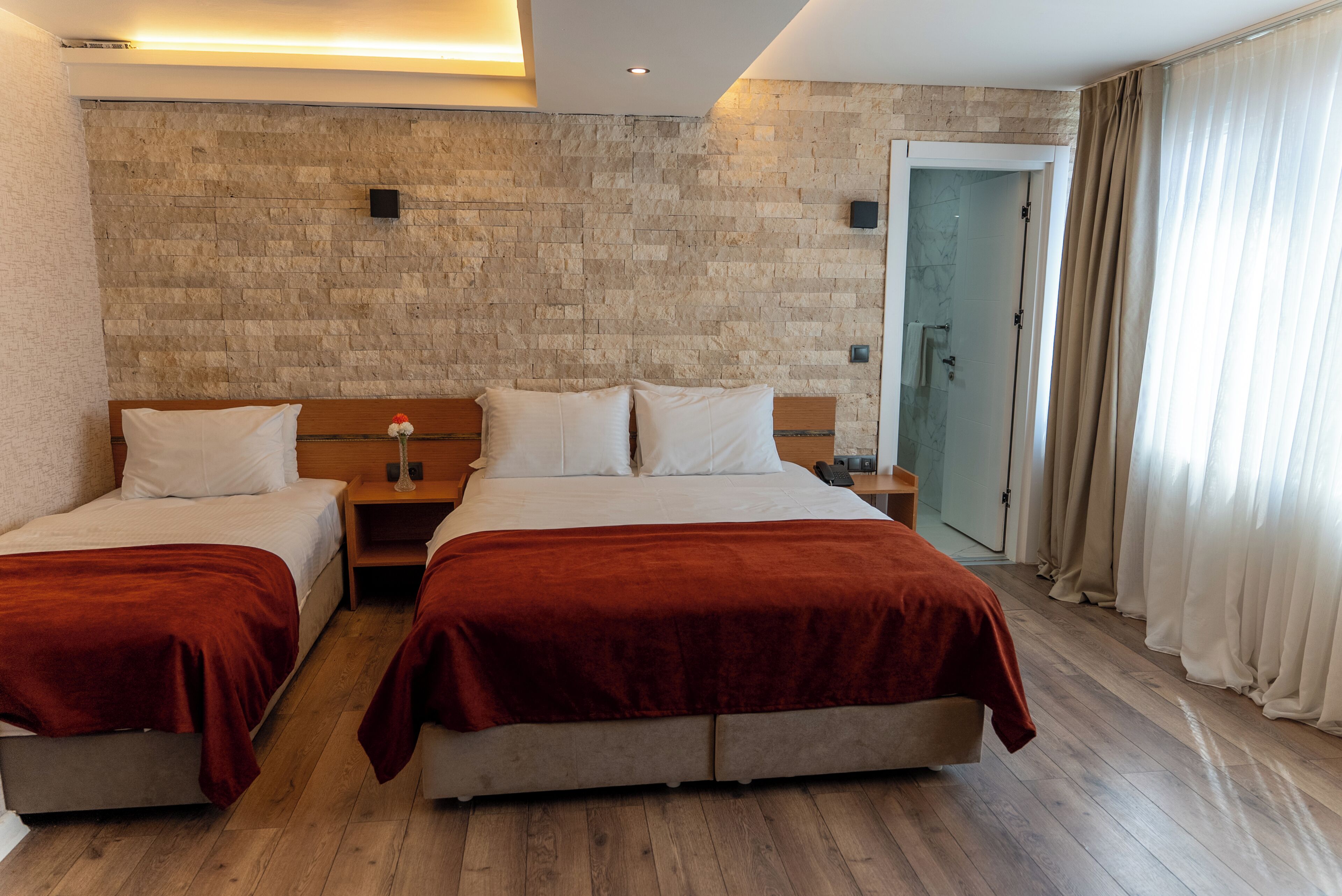 A hotel room with two beds, a wooden floor, a stone wall, and a door leading to a bathroom.