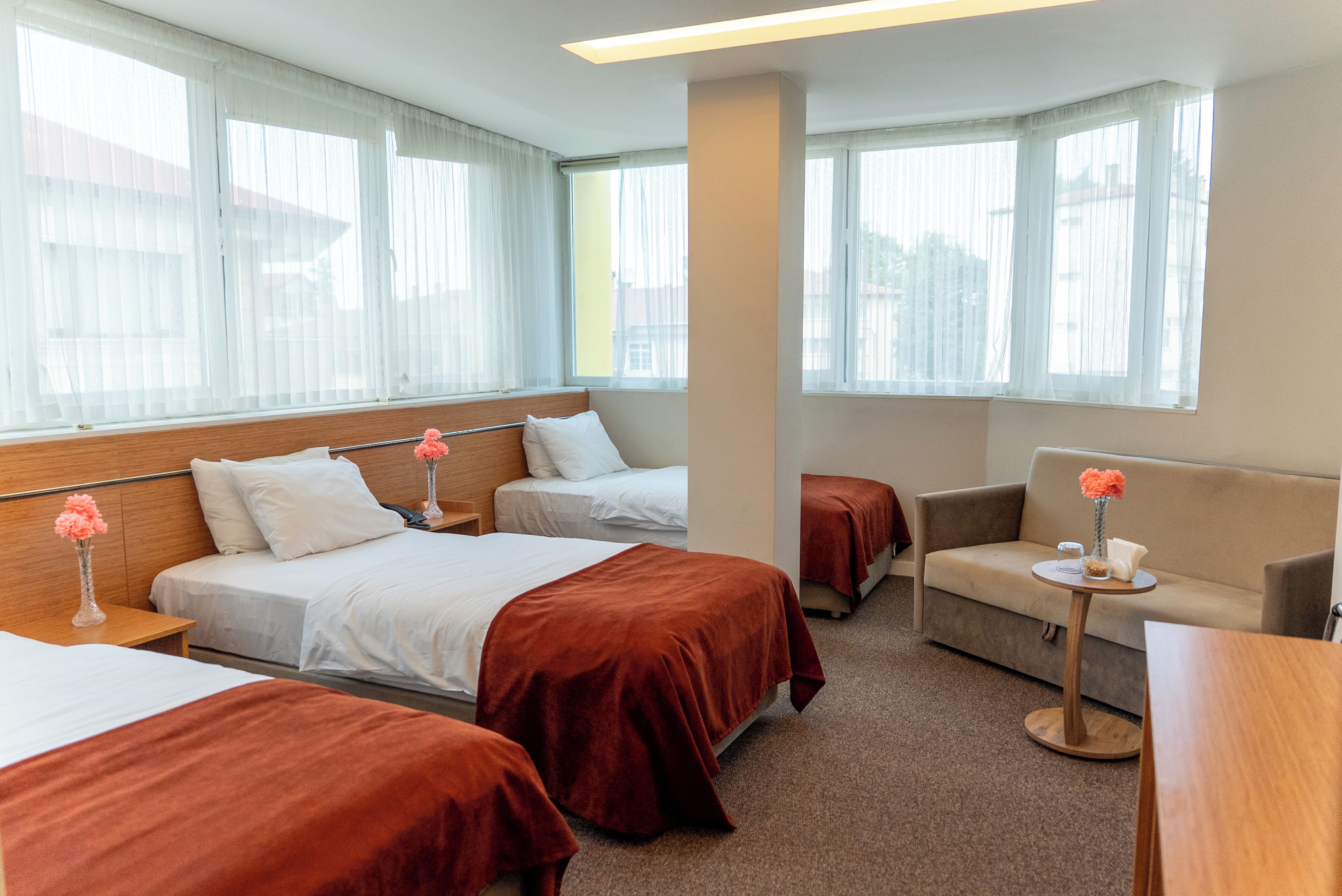 A hotel room with two beds, a sofa, a small table, and large windows with sheer curtains.