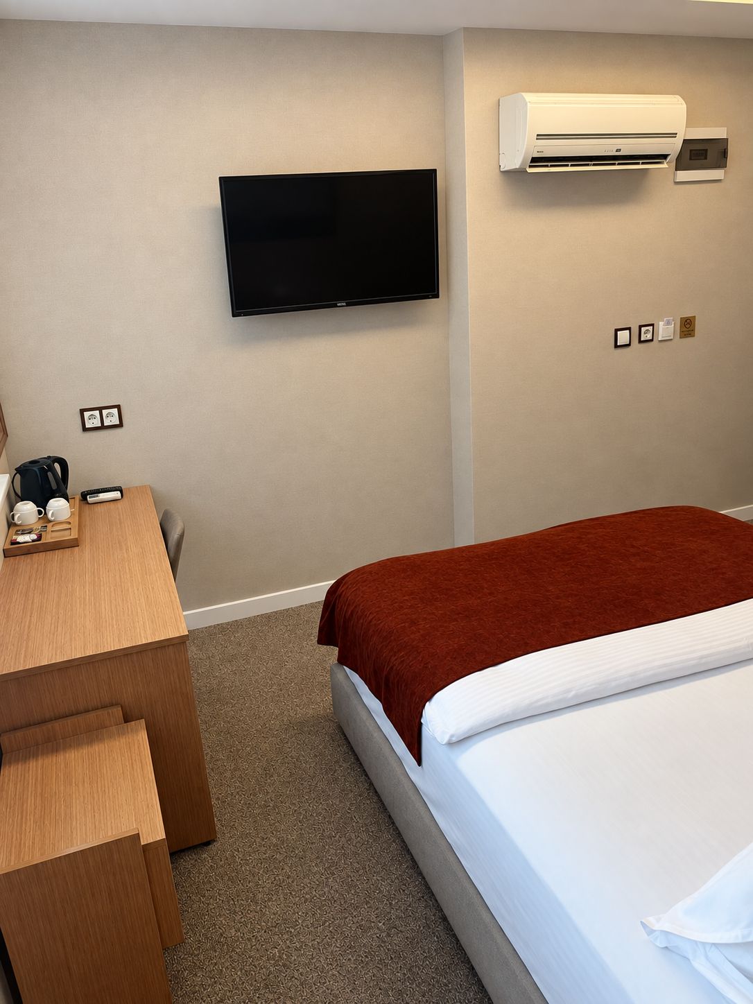 A hotel room with a bed, a desk with a coffee maker, a television, and an air conditioning unit.