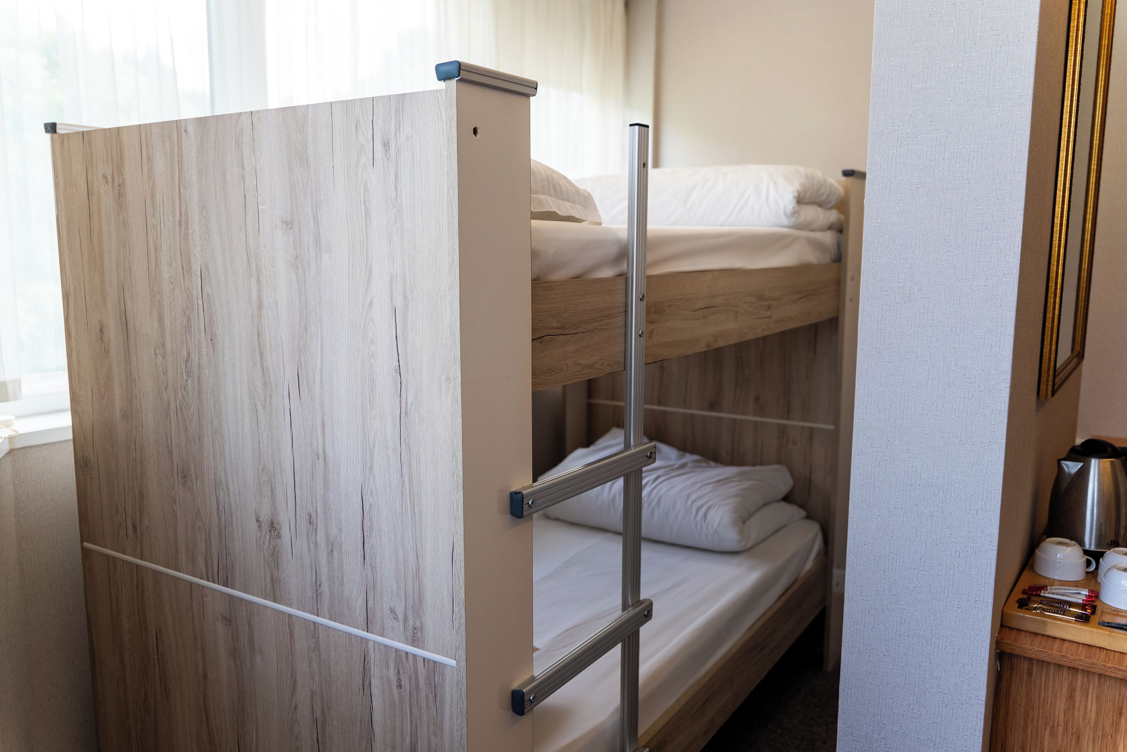 Ein Etagenbett mit Holzkopfteil, eine untere Schlafstelle mit Matratze und Kissen sowie ein Nachttisch mit Wasserkocher und Tassen.