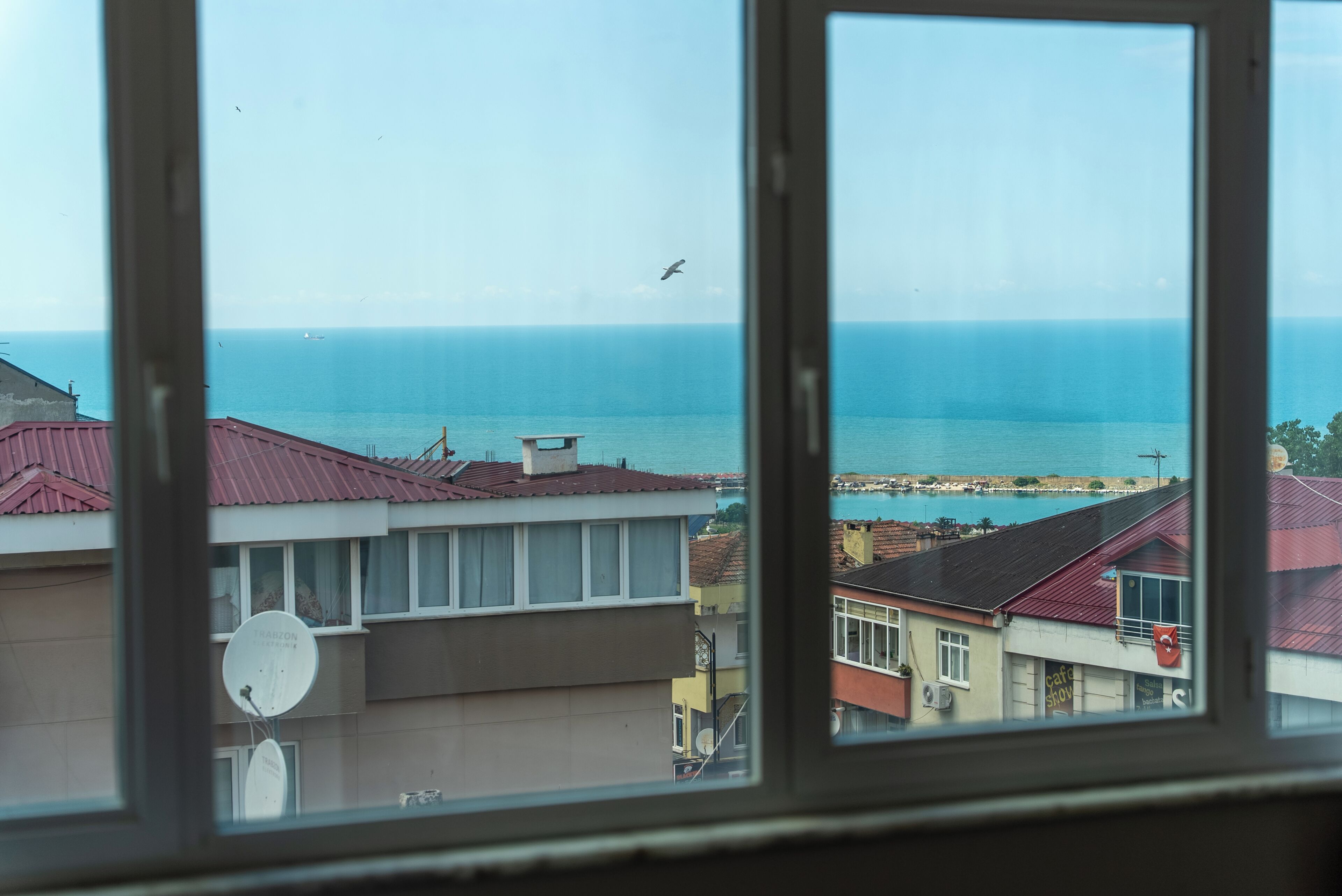 Vista a través de una ventana mostrando una zona costera con edificios, cielo despejado y un barco a lo lejos.