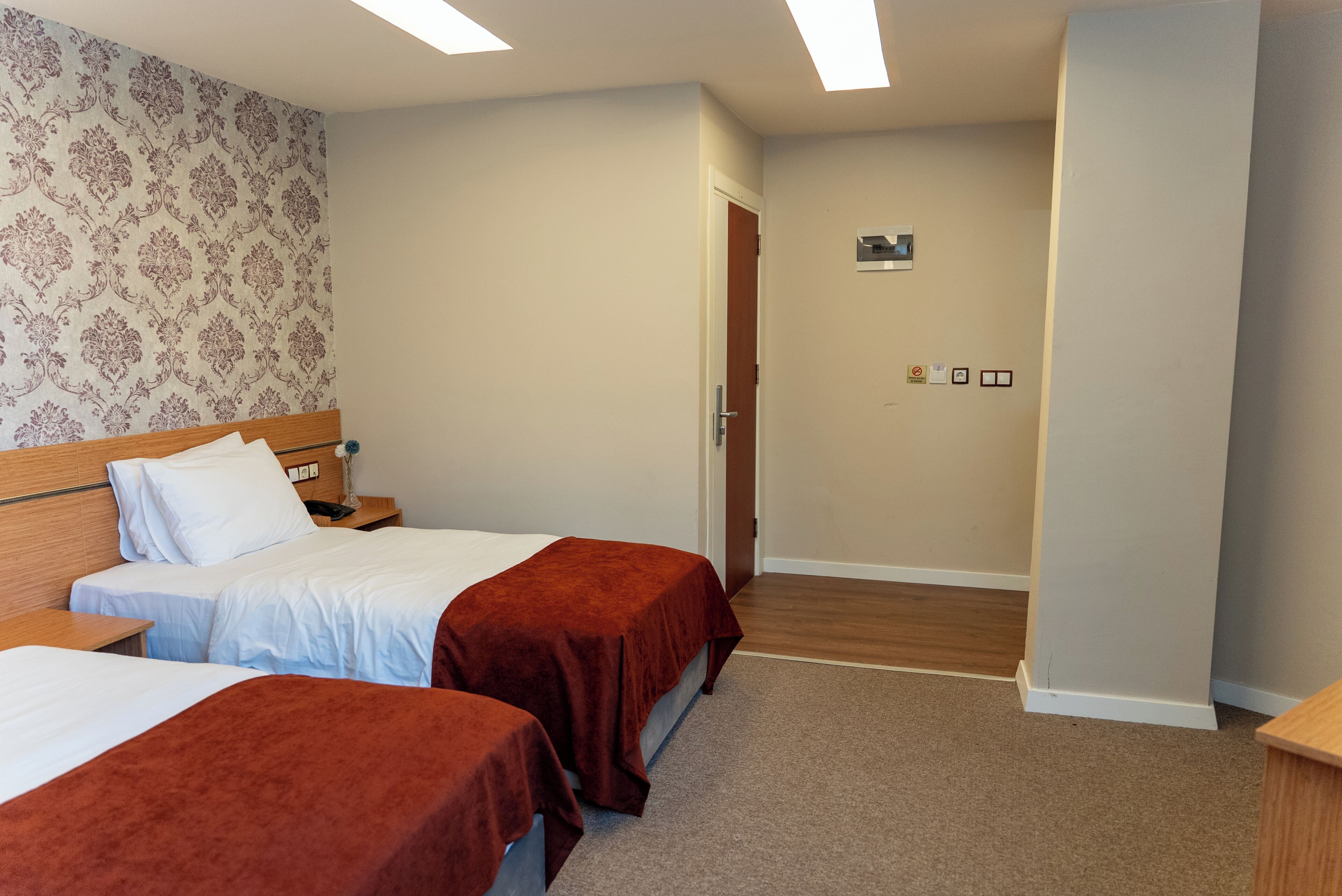 A hotel room with two beds, a wooden headboard, a patterned wallpaper, a carpeted floor, and a door leading to another room.