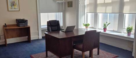 Una sala de conferencias con una mesa de madera oscura, dos sillas, una computadora portátil y una impresora sobre una mesa pequeña. Hay luces colgantes, una ventana con persianas y una planta en el alféizar.