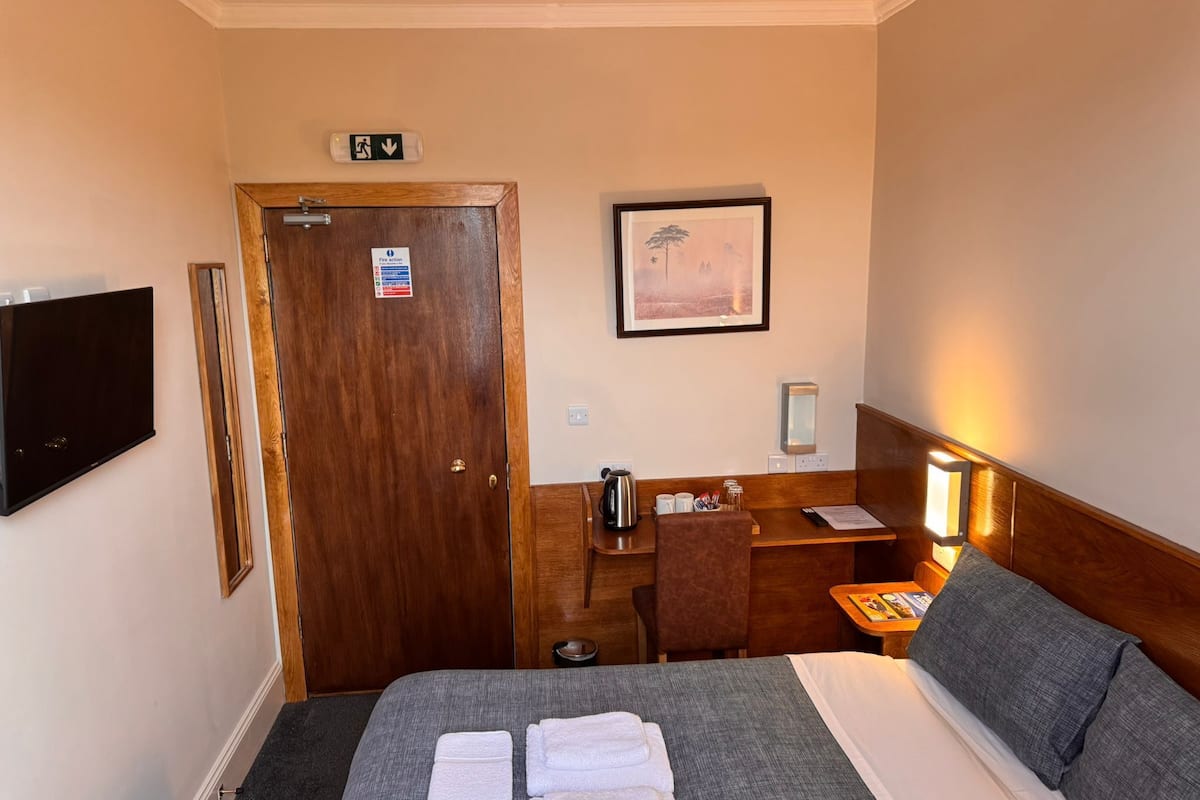 A hotel room with a bed, a desk, a chair, a TV, a framed picture, and a nightstand with a lamp.