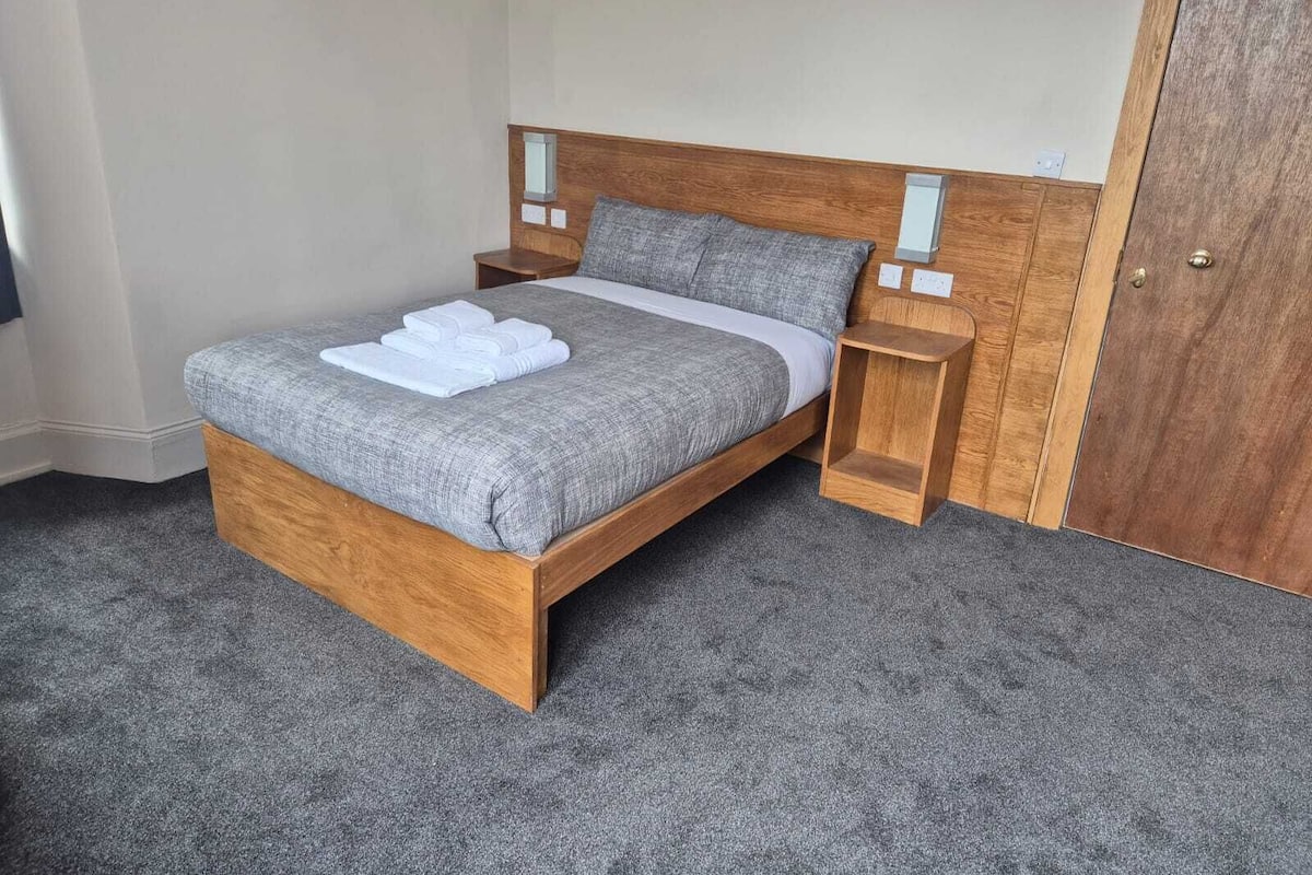 A neatly made bed with a grey duvet and white towels, wooden headboard, bedside tables, and a wooden door.