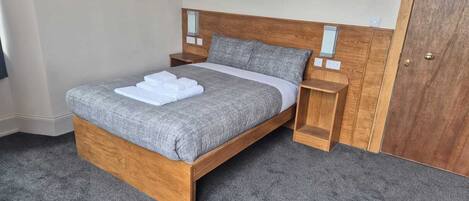 A neatly made bed with a grey duvet and white towels, wooden headboard, bedside tables, and a wooden door.