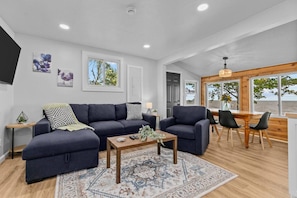 A modern living room with a dark blue sofa, a wooden coffee table, and a patterned rug. There is a dining area with a wooden table and chairs, and large windows offering a view of the outdoors. - New! Lake Front|PrivateDock|Kayaks&Paddleboards! (Chilton)