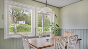 A dining area with a wooden table and white chairs, large windows with a view of greenery, and a hanging light fixture.