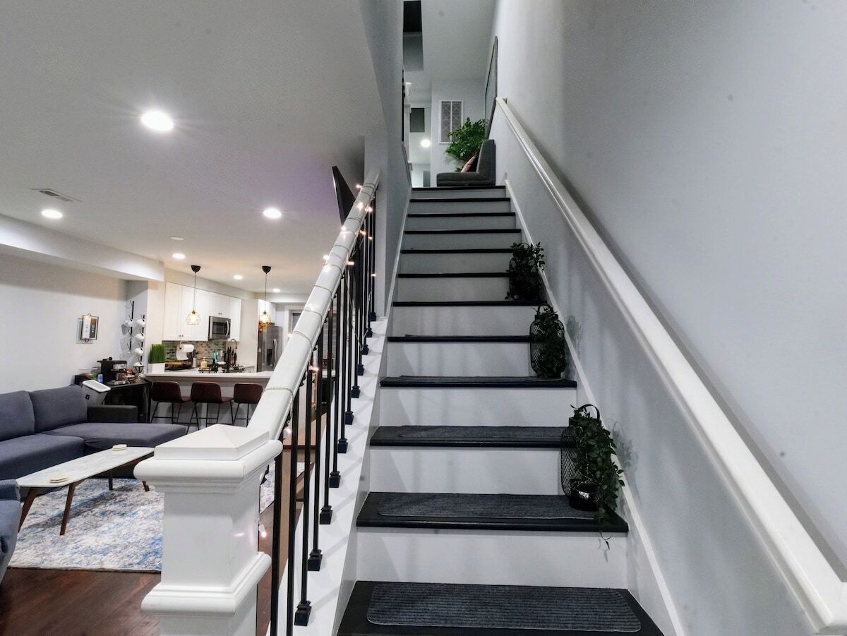 Una escalera con barandilla blanca, una sala de estar con sofá y mesa de centro, y una cocina al fondo.