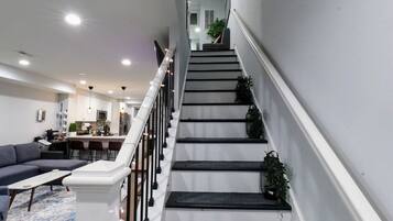 A staircase with a white railing, a living room with a sofa and coffee table, and a kitchen in the background.