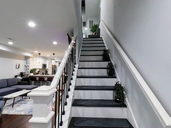 A staircase with a white railing, a living room with a sofa and coffee table, and a kitchen in the background. - Modern 2BR, 2.5BA home in the heart of NoMa/Trinidad (Washington)