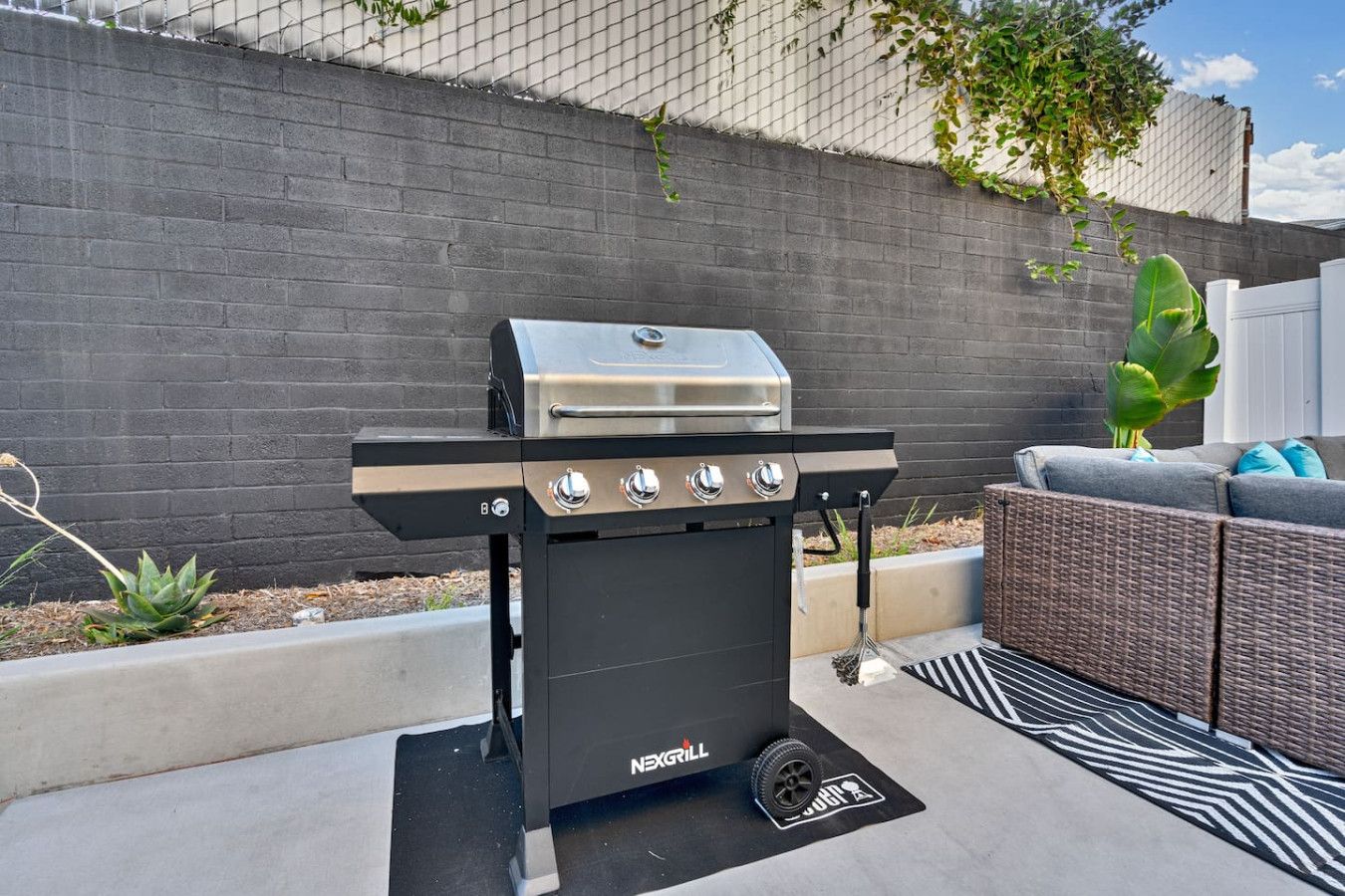 A black and silver grill with four burners, positioned on a concrete patio with a black and white patterned rug.