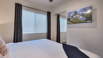 A hotel room with a bed, a painting of a mountain landscape, and a window with blinds.
