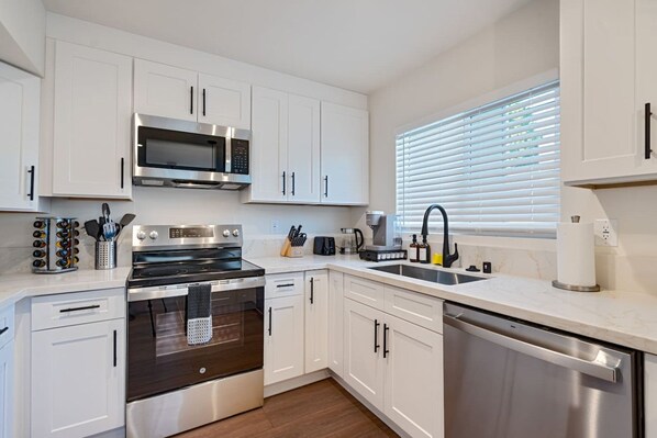A modern kitchen with white cabinets, stainless steel appliances, a dishwasher, and a window with blinds.