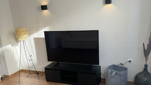 Een moderne woonkamer met een flatscreen televisie op een zwarte standaard, een vloerlamp en een vaas met gedroogde planten.