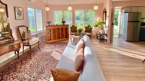A spacious living room with a sofa, chairs, a wooden console table, and a view of the outdoors through large windows. - Dream house by a Pond, 3-bed home in Woodridge NY w/ WiFi, AC, embraced by trees (Woodridge)