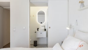 Et hotelværelse med en seng, et natbord, en lampe, et spejl, en vask og en skraldespand.