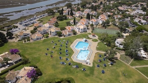 Ovenudblik over et boligområde med en stor swimmingpool omgivet af huse, en tennisbane og en parkeringsplads.