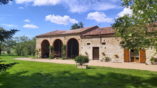A stone building with three arched doorways, a tiled roof, and a paved driveway.