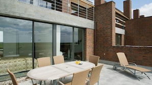An outdoor patio with a round table and wicker chairs, a lounge chair, and a glass wall reflecting the sky. - 4 Person Holiday Home on a Holiday Park in Romo (Rømø)