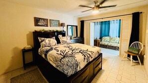 Une chambre à coucher comprenant un lit, une table de chevet, un ventilateur de plafond, une fenêtre avec des rideaux et une chaise.