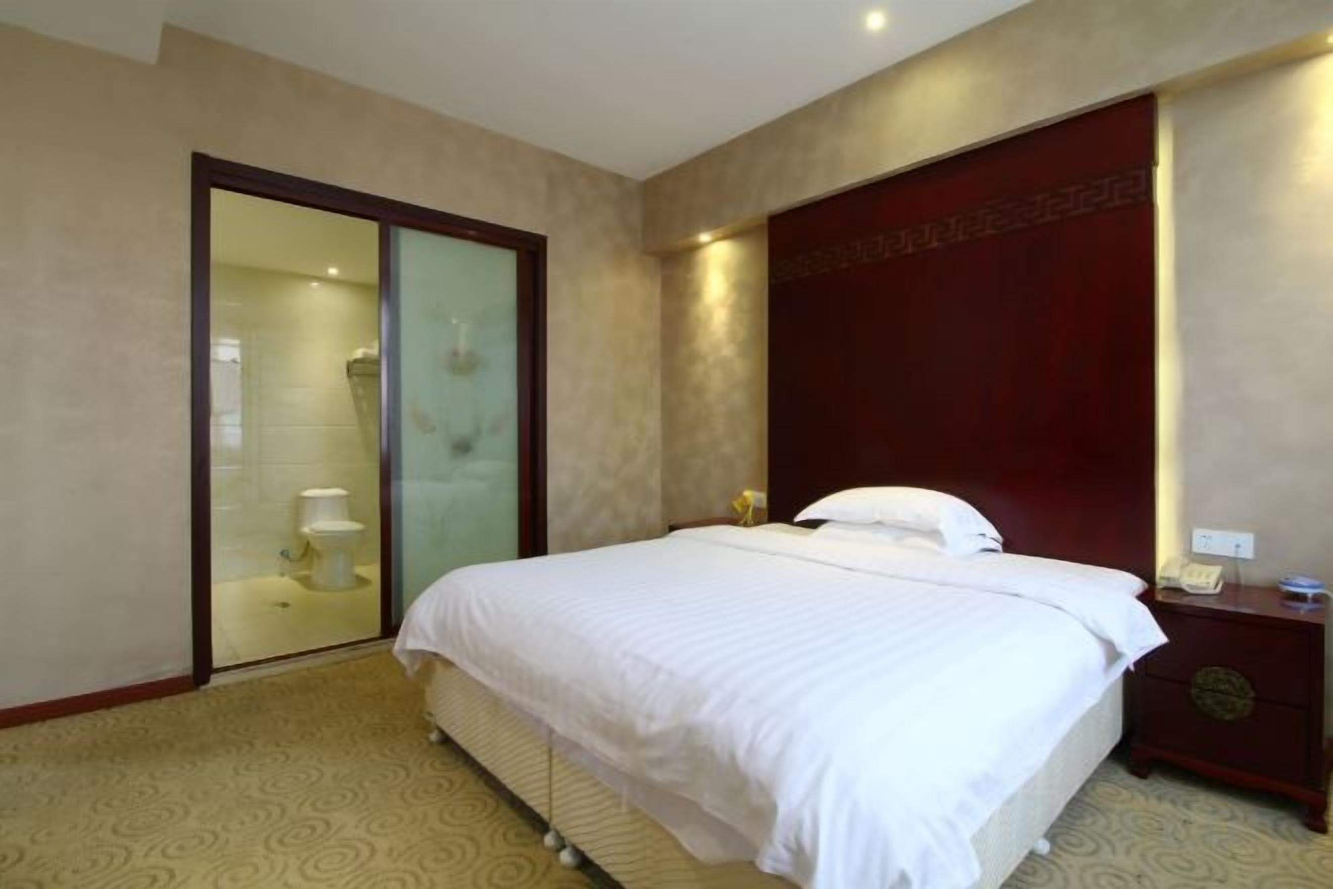 A hotel room with a bed, a nightstand, a wooden headboard, a glass shower enclosure, and a carpeted floor.