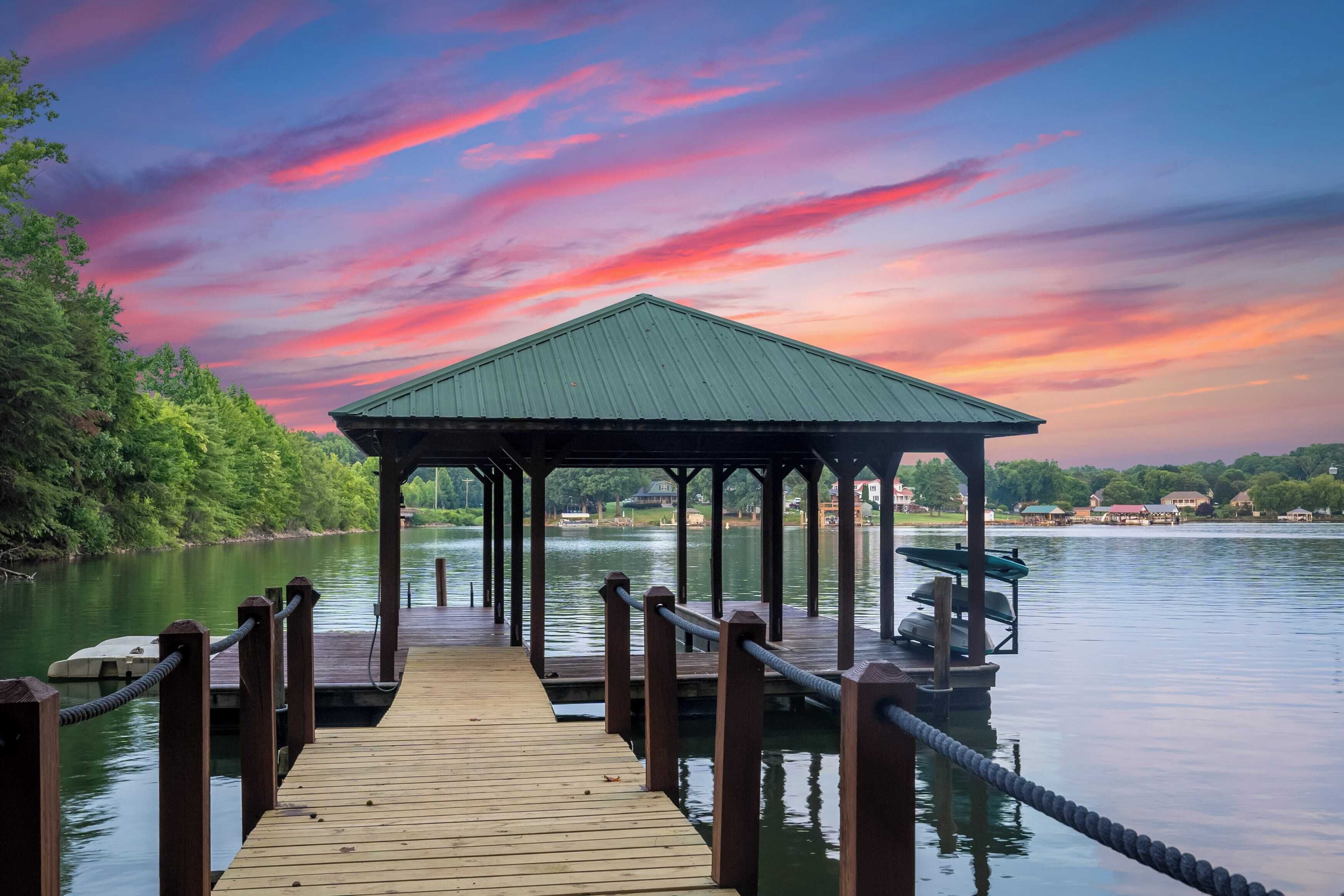 A wooden pier leading to a gazebo with a green roof, overlooking a calm lake with a sunset sky.