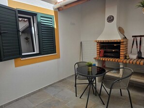 Un patio con horno de ladrillo, dos sillas, una mesa y una ventana con contraventanas.