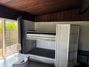 Uma cama de beliche com estrutura metálica e colchão, uma pequena mesa com luminária, e uma janela com vista para o verde.