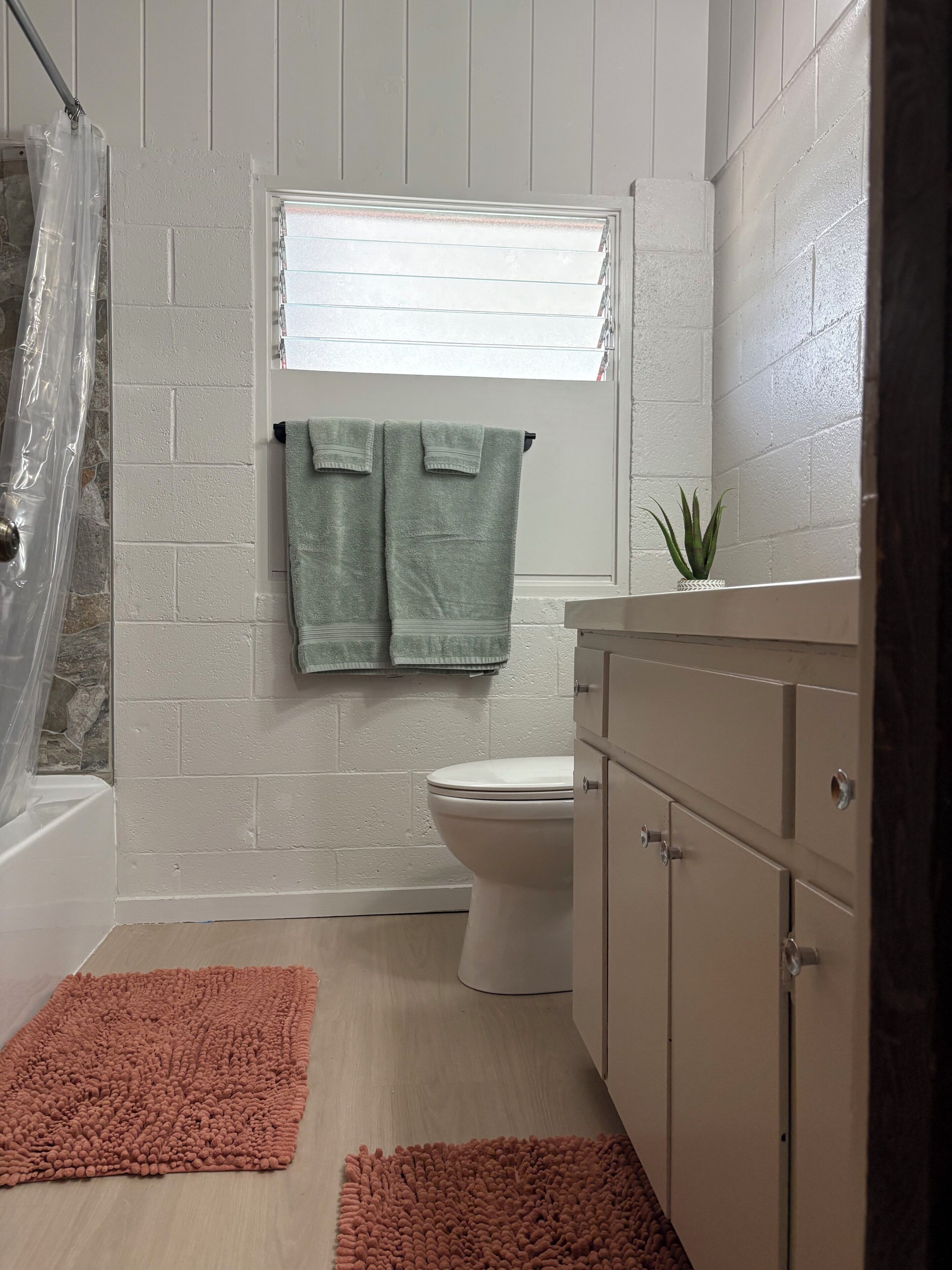 Un baño con pared de azulejos blancos, inodoro, ventana y una toalla colgada, además de una pequeña planta en el mostrador.