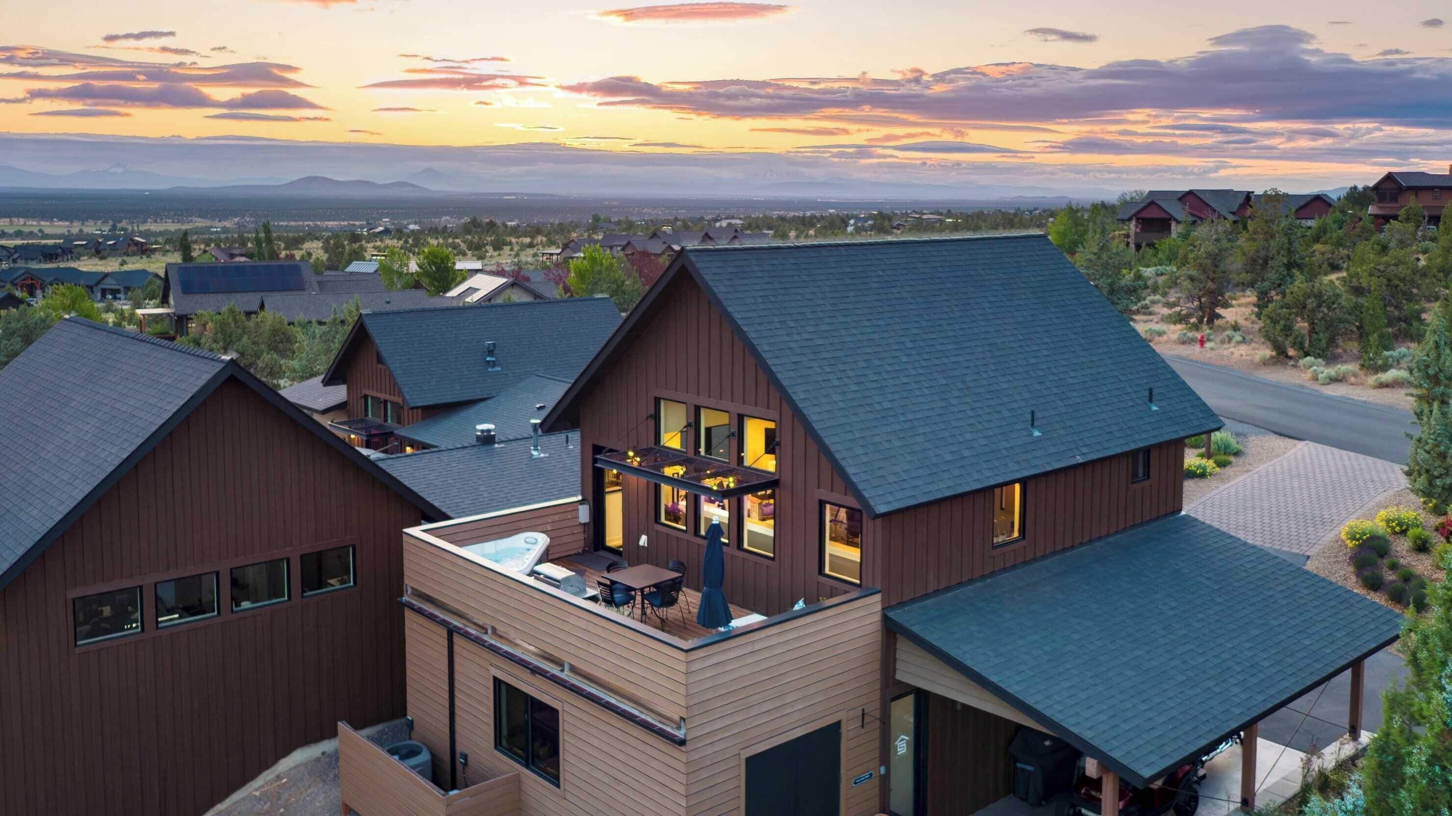 A modern multi-story house with a large balcony and a hot tub, surrounded by a suburban landscape with mountains in the background.