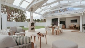Un patio exterior con una pérgola blanca, un estufa de leña y una zona de comedor con sillas y mesas.
